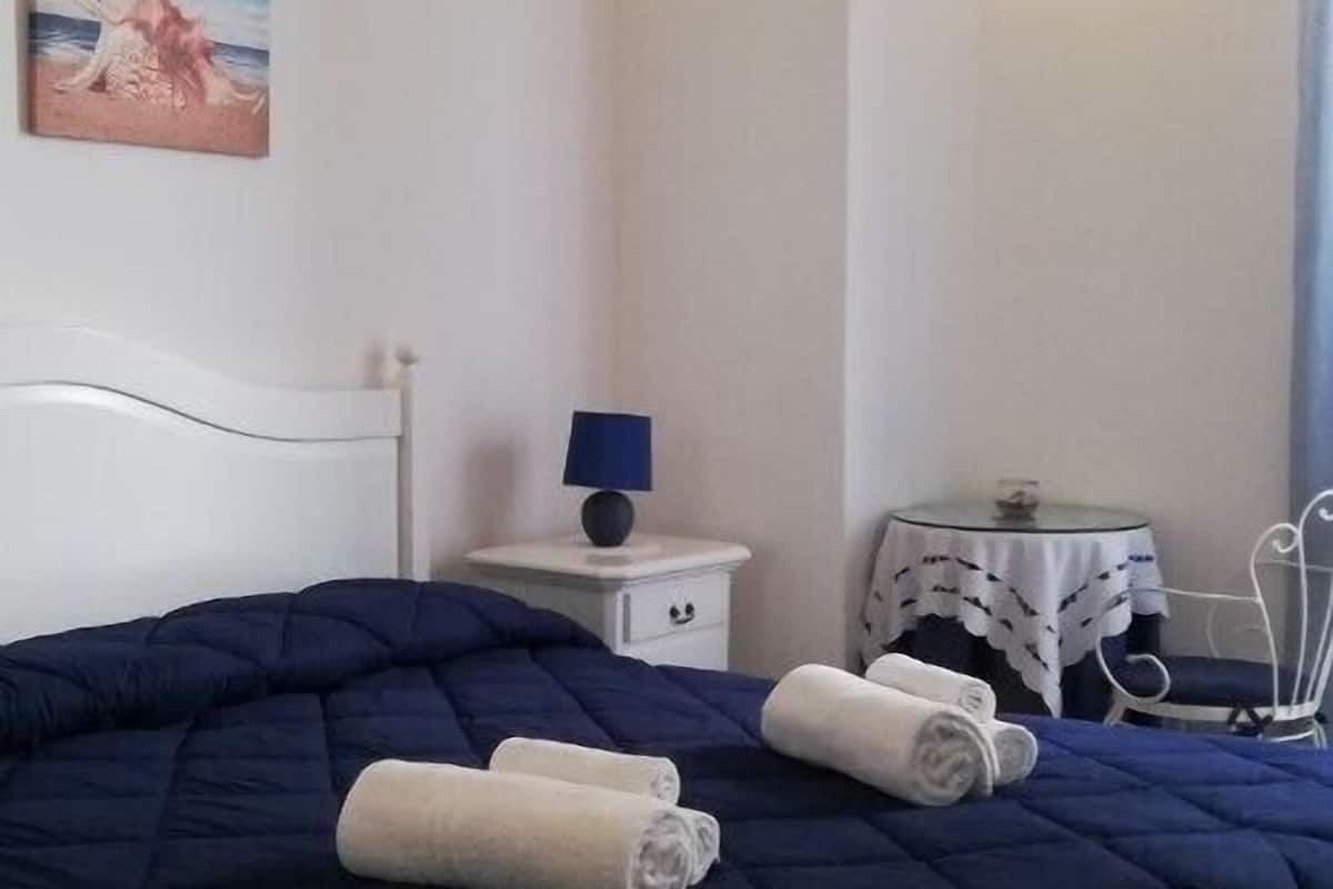Una cama bien hecha con una colcha azul oscuro y toallas enrolladas, una mesita de noche con una lámpara, una pequeña mesa con un adorno y un aire acondicionado de pared.