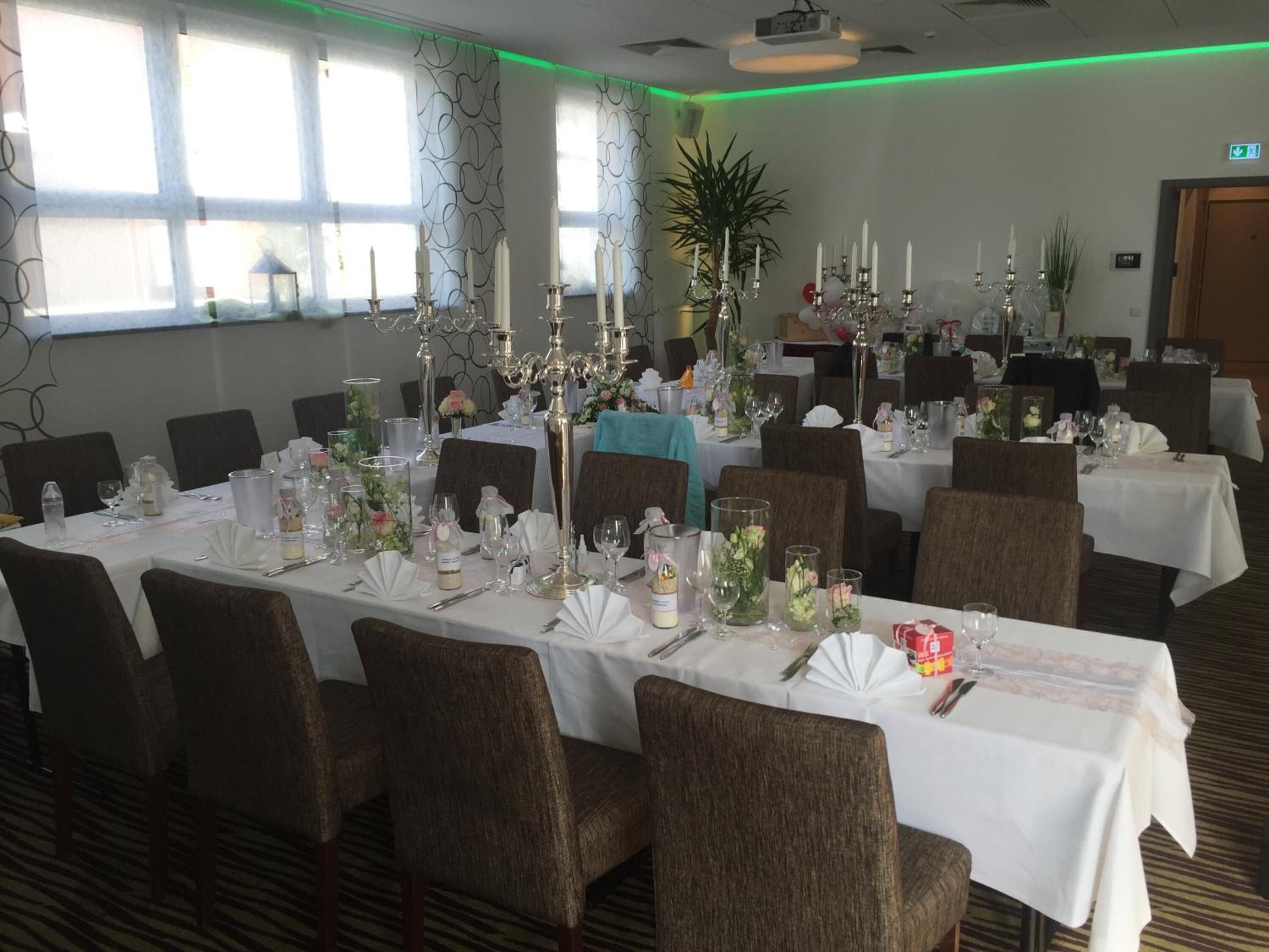 A banquet hall with multiple tables set for dining, featuring white tablecloths, floral centerpieces, and candle holders.