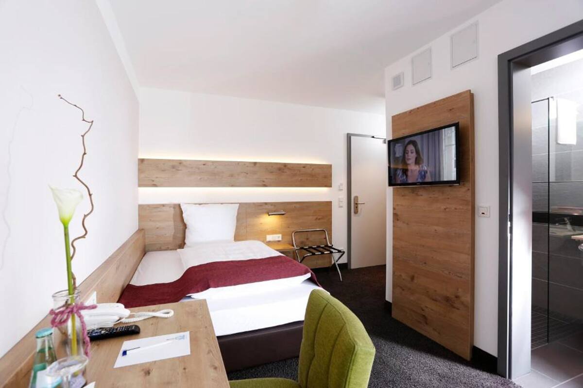 Een moderne hotelkamer met een bed, een bureau met een vaas en afstandsbedieningen, een televisie aan de muur en een houten hoofdbord.