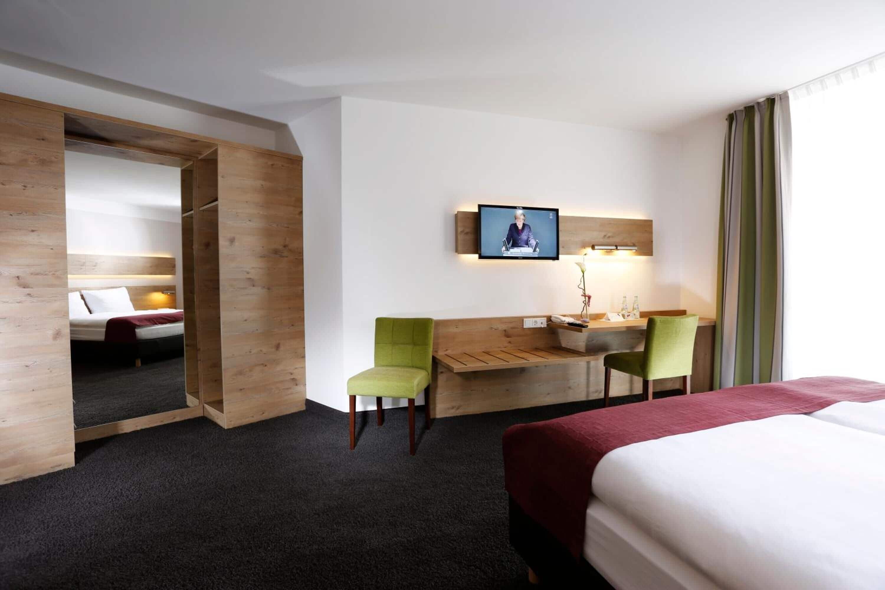 A hotel room with a bed, a desk with two chairs, a television on the wall, and a wooden wardrobe.