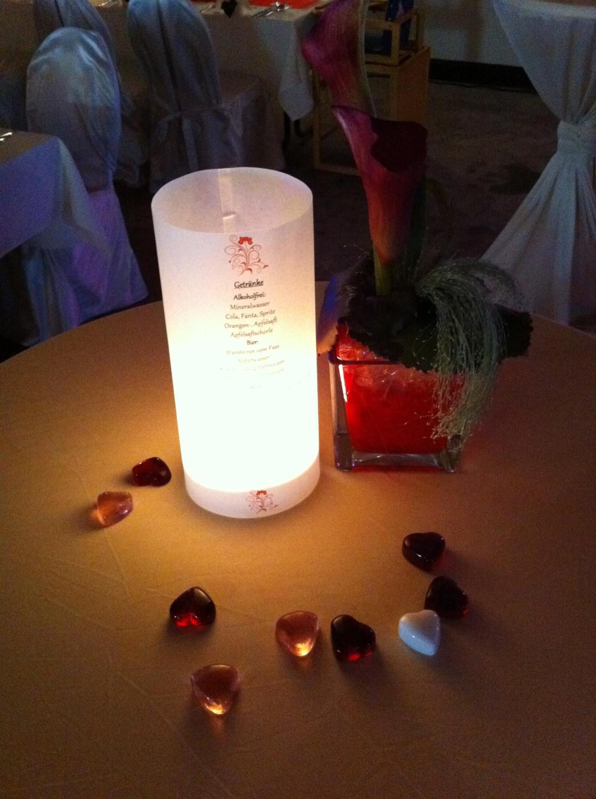 A table with a lit candle, a small red vase with a flower, and scattered heart-shaped stones.