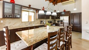 A kitchen with granite countertops, dark wooden cabinets, a central island with chairs, and pendant lighting. - Grand Laguna Beach Properties by Calisto Stays (Sosúa)