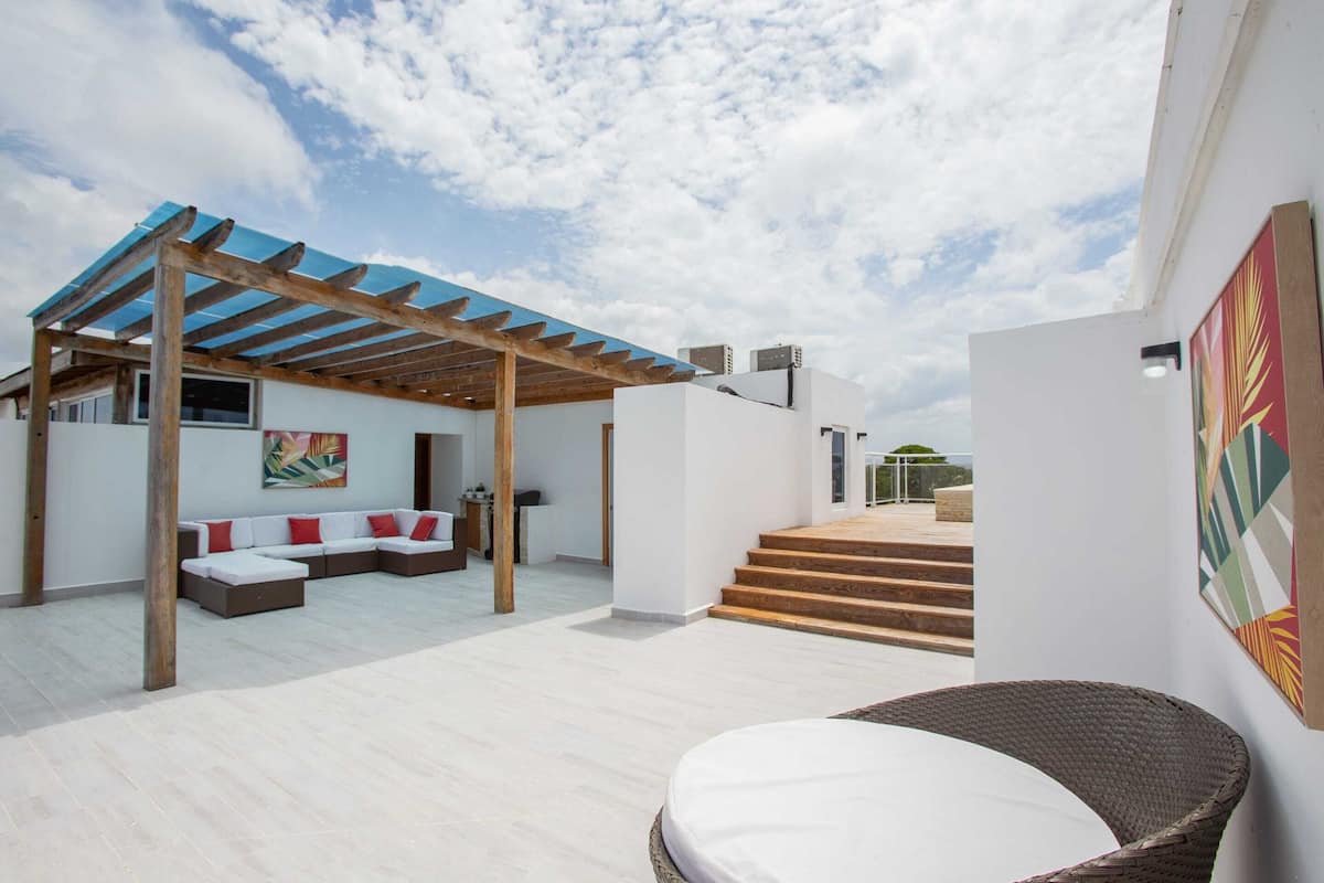 Un espace de vie extérieur moderne, doté d’un patio couvert, d’un coin salon avec des coussins et d’un fauteuil en osier.