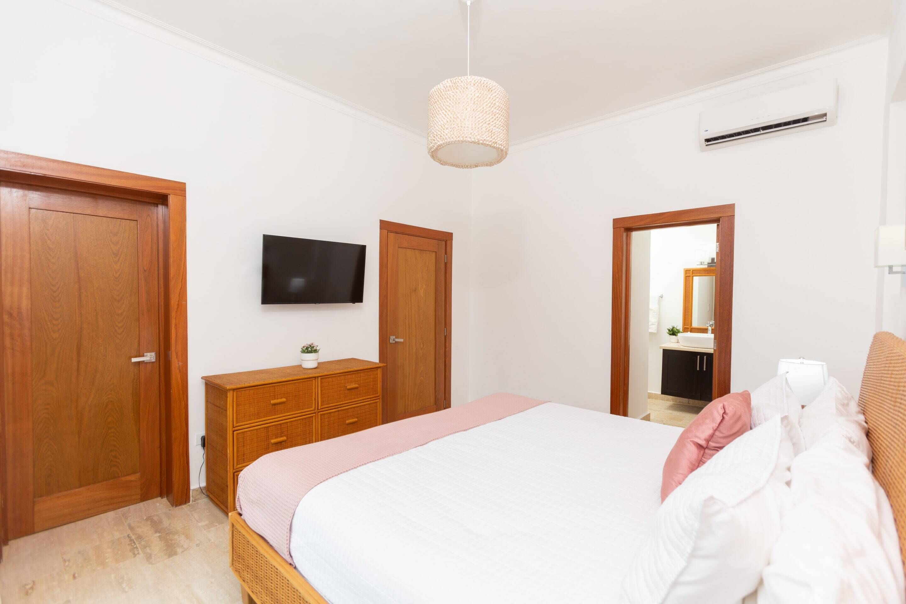 A bedroom with a bed, wooden wardrobe, a television, and a bathroom visible through an open door.