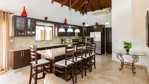 A spacious kitchen with dark wooden cabinets, a granite countertop, and a central island with bar stools. There is a dining area with a glass table and chairs, and a small round table with a plant. - Grand Laguna Beach Properties by Calisto Stays (Sosúa)