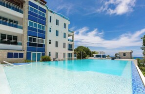 A modern hotel with a large swimming pool, multiple balconies, and a clear view of the ocean.