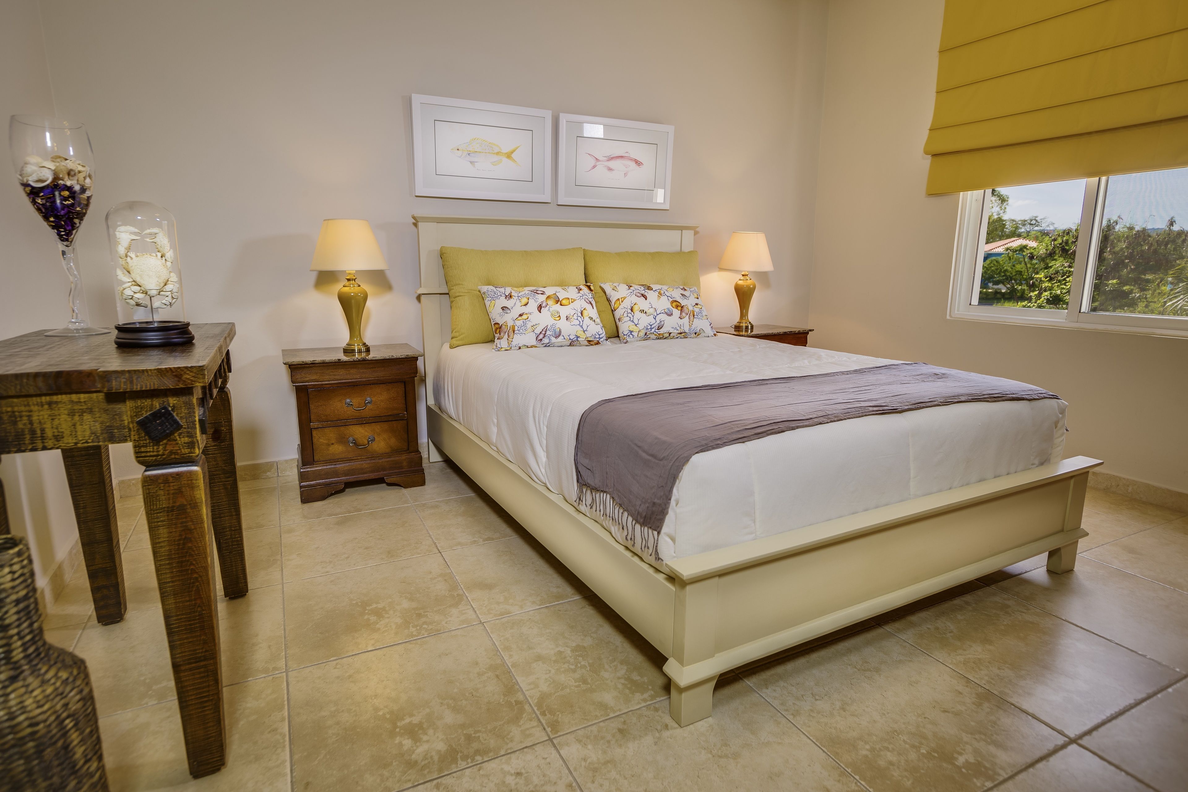 A bedroom with a bed, two bedside tables with lamps, a nightstand, a window with a view, and two framed pictures on the wall.