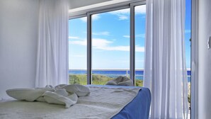 Ein Schlafzimmer mit großem Fenster, weißen Vorhängen, einem Bett mit blauer Matratze und Blick aufs Meer.