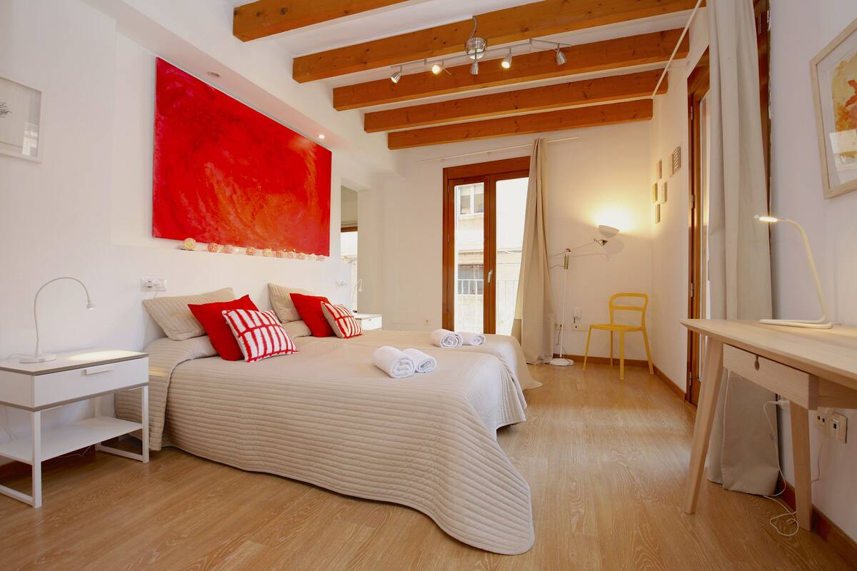 Ein Schlafzimmer mit einem großen Bett, einem Schreibtisch und einem Stuhl. An der Wand hängt ein rotes Gemälde und ein Fenster mit Vorhängen.