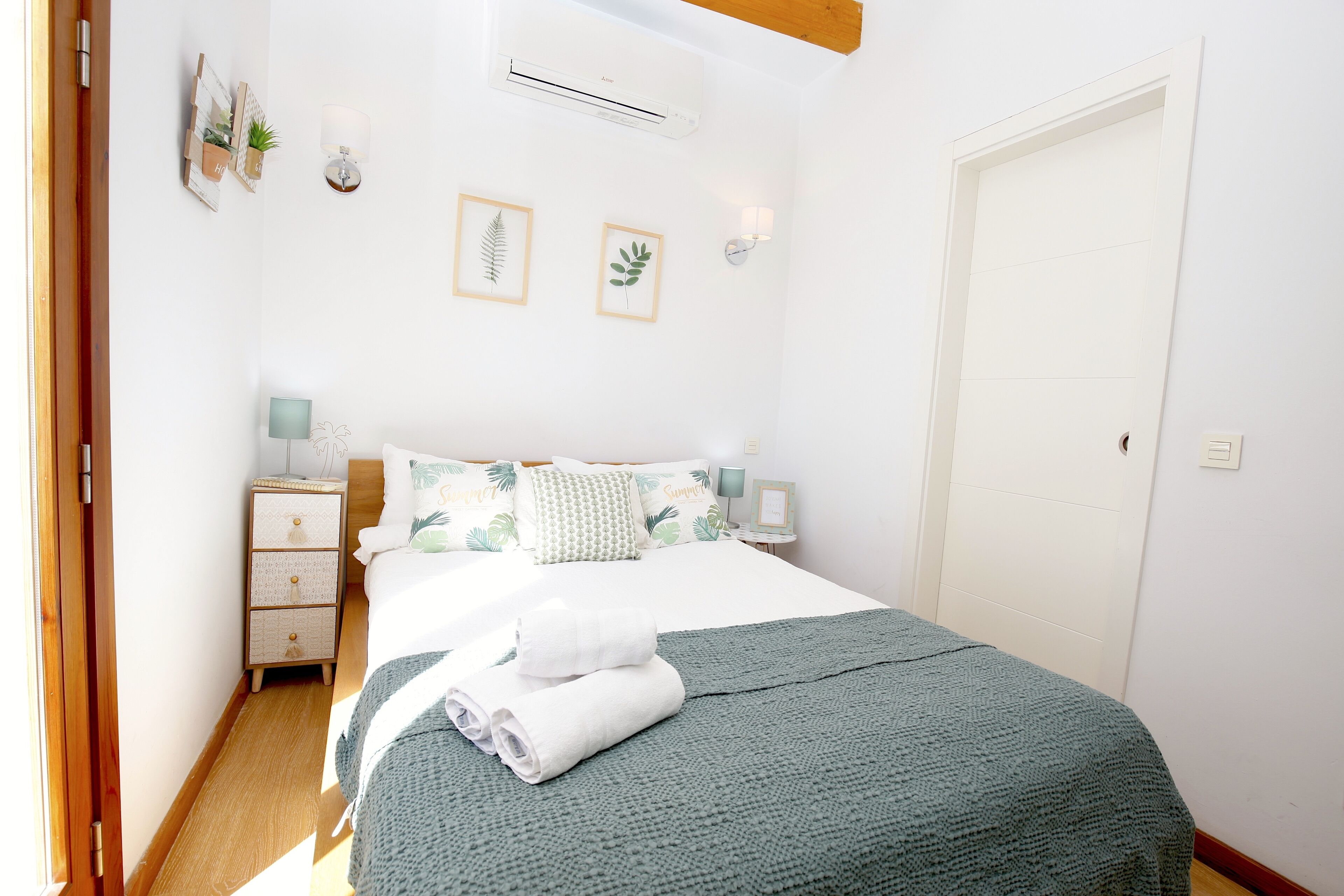 A neatly made bed with a green blanket, a bedside table with a lamp, and a wall-mounted air conditioner.