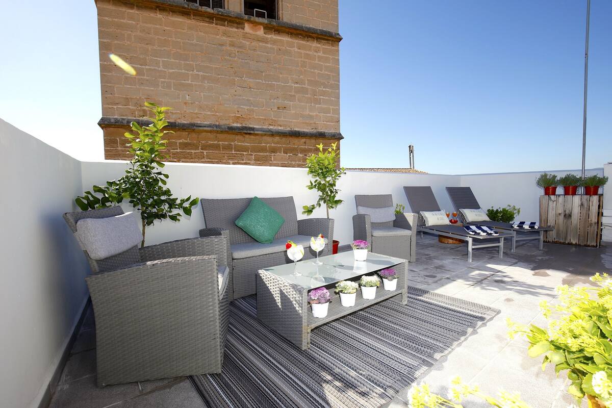 Eine Dachterrasse mit Korbmöbeln, einem Glastisch und Topfpflanzen.
