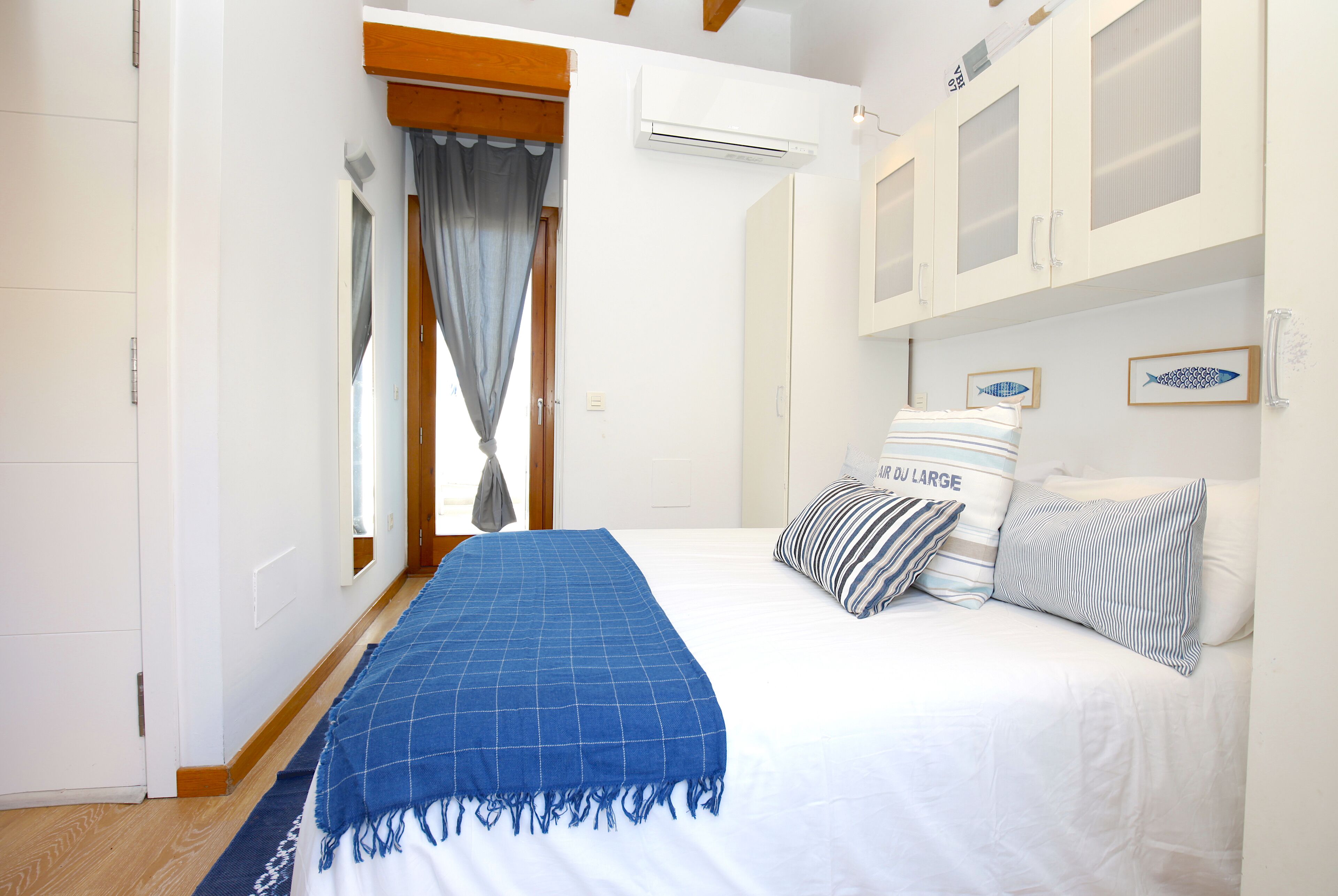 A bedroom with a bed, a blue blanket, and striped pillows. There is a window with curtains, an air conditioning unit, and a wardrobe with glass doors.