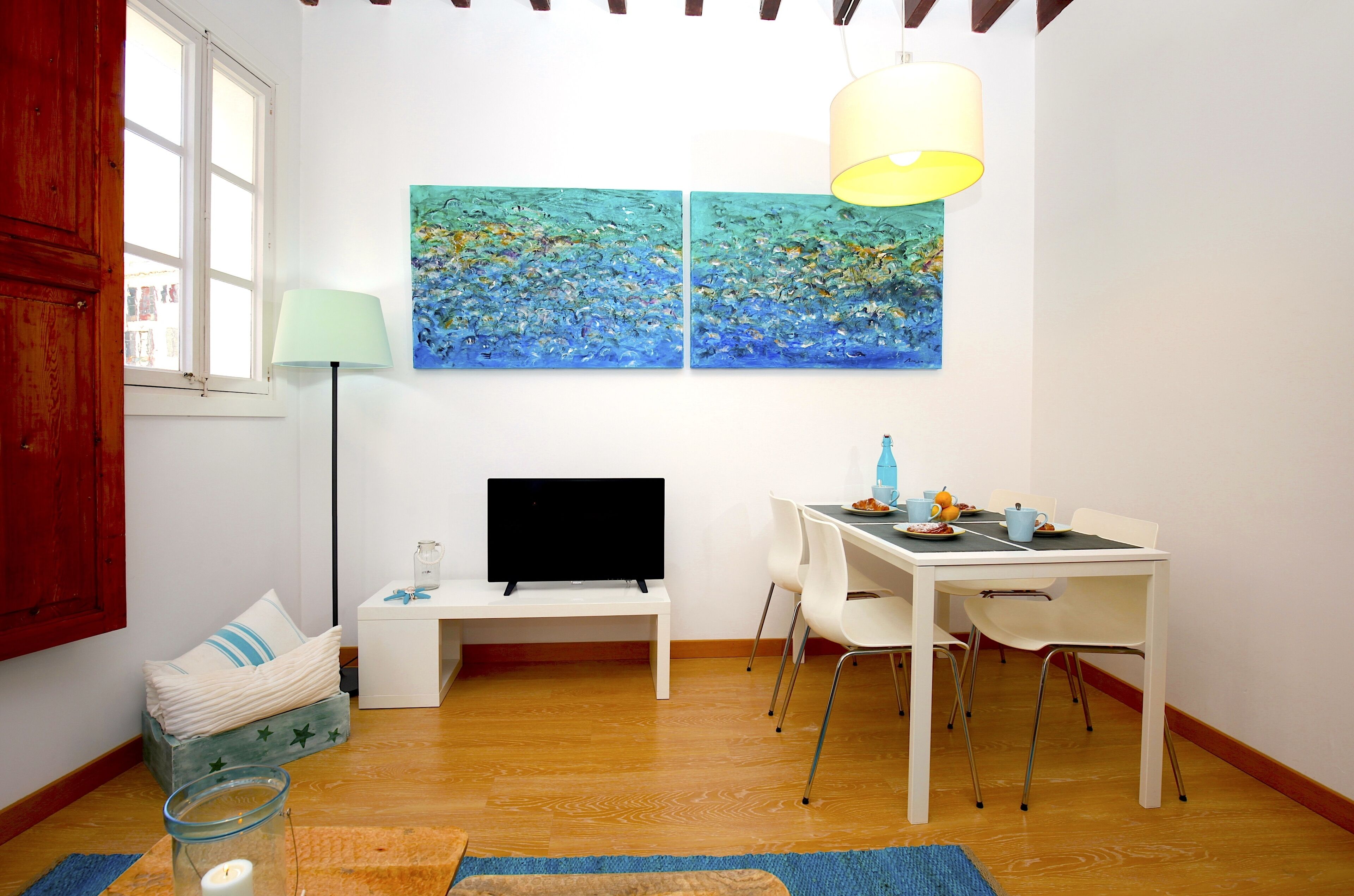 A room with a wooden floor, white furniture, a TV on a stand, and two paintings on the wall.