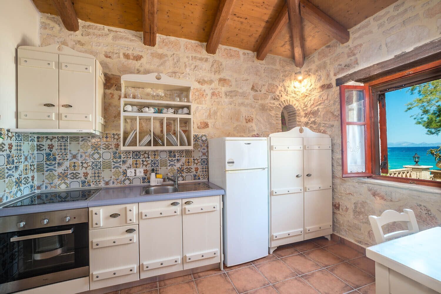 Une cuisine de taille réduite avec un mur en pierre, des poutres en bois et une vue sur la mer par une fenêtre.