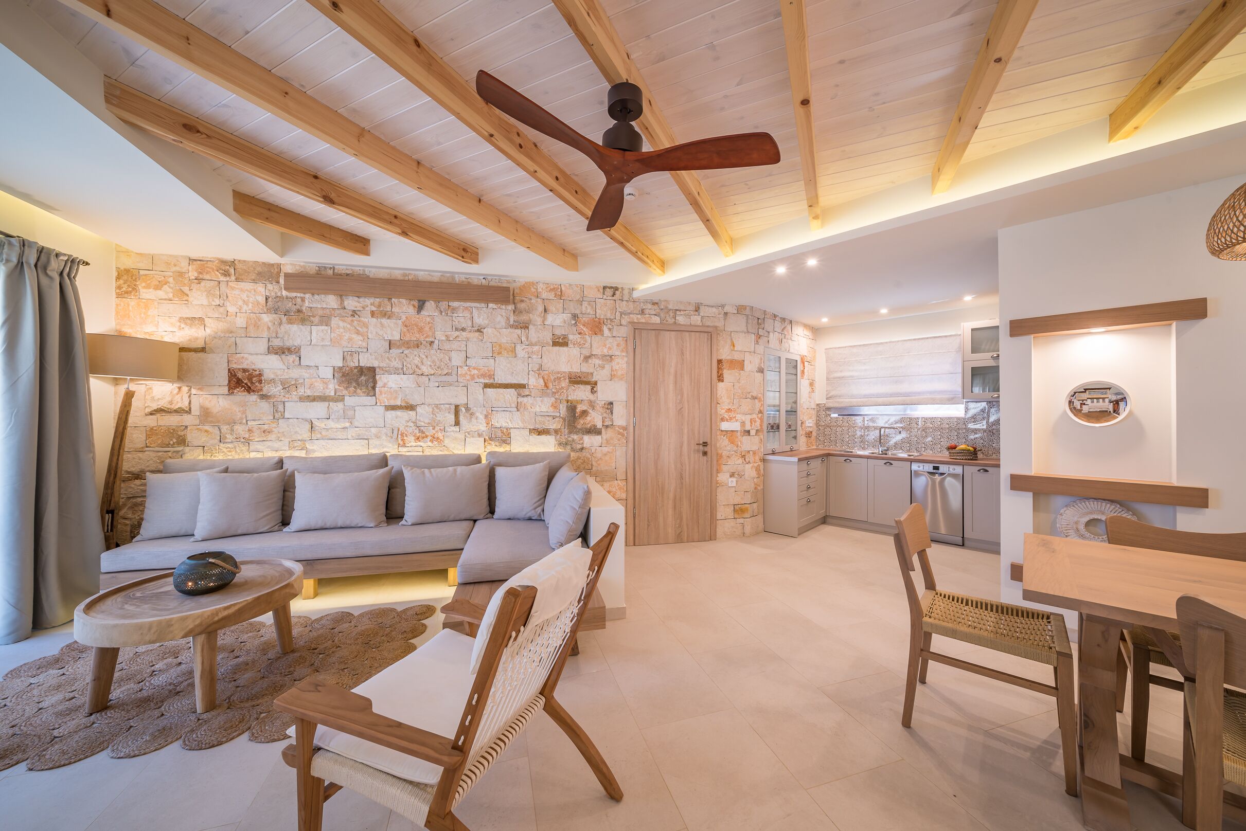 A modern living room with a stone wall, wooden ceiling beams, a sofa, a dining table, and a kitchen in the background.