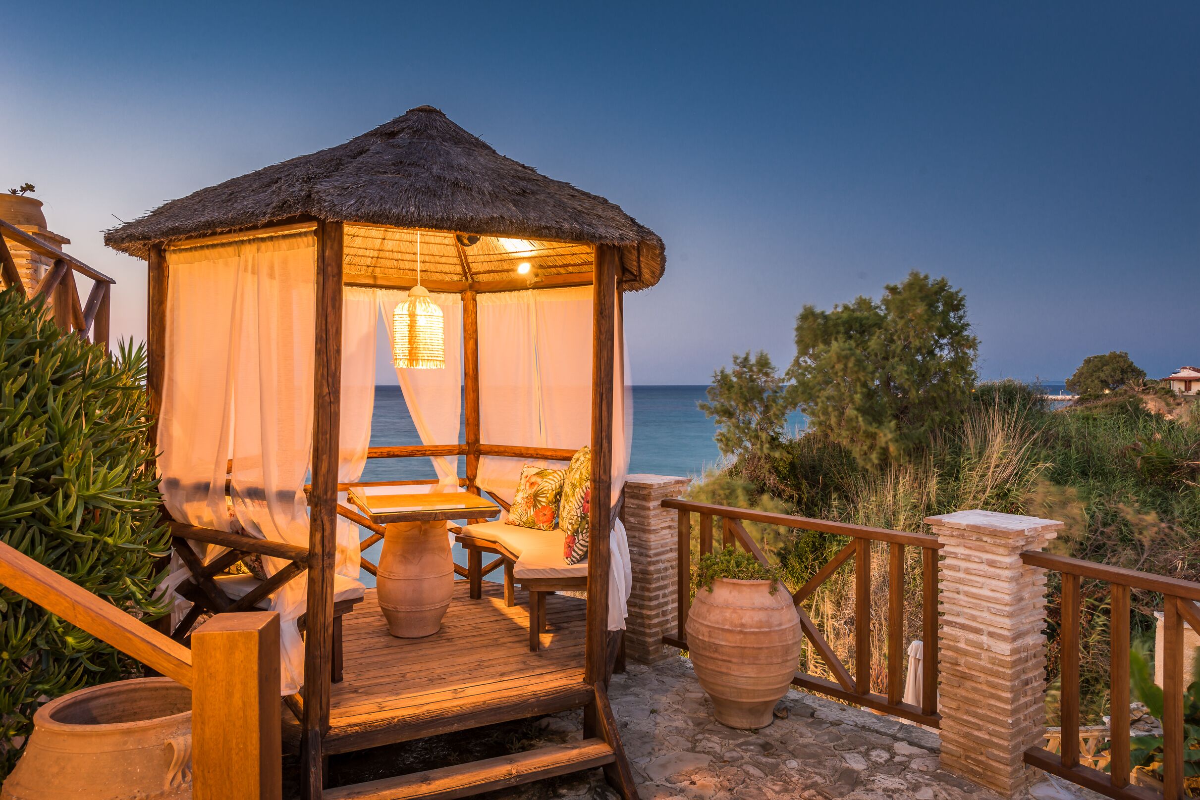 A wooden deck with a thatched-roof gazebo, a stone pillar, and a view of the sea.