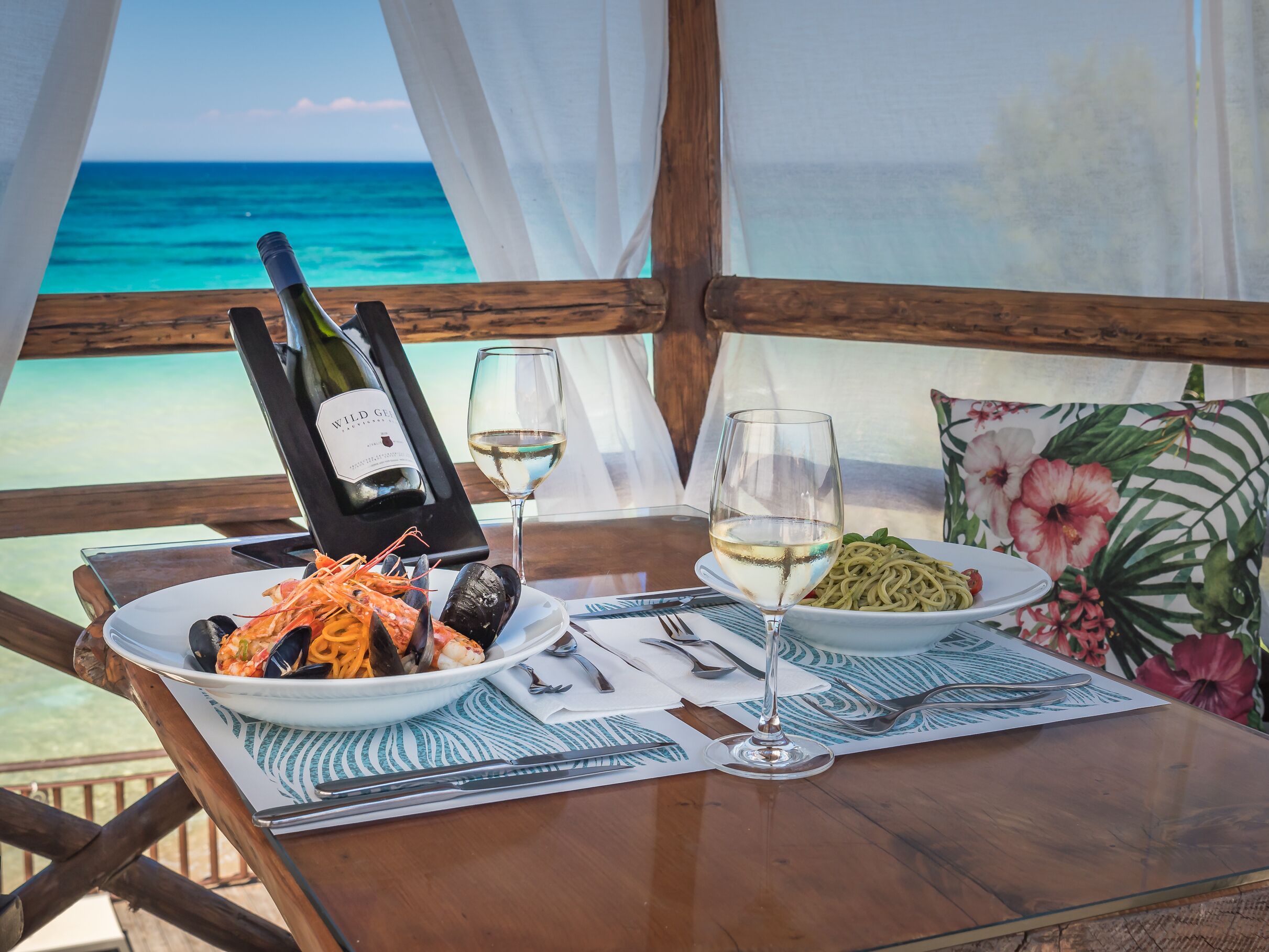 Une table dressée avec des assiettes de fruits de mer et de pâtes, une bouteille de vin et un verre de vin, le tout sur fond de plage et de vue sur l’océan.