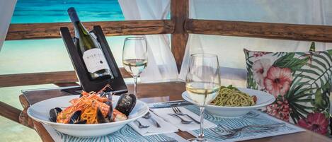 Une table dressée avec des assiettes de fruits de mer et de pâtes, une bouteille de vin et un verre de vin, le tout sur fond de plage et de vue sur l’océan.