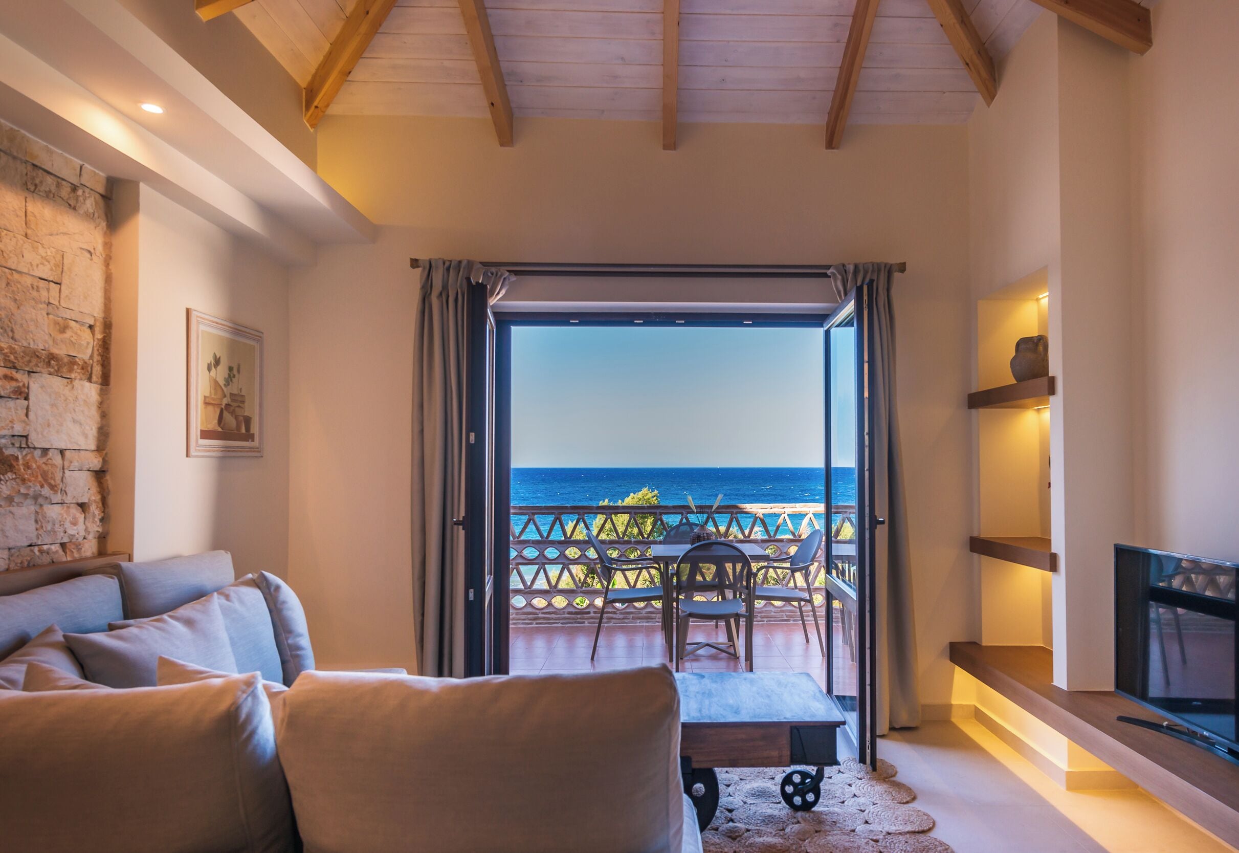 A cozy living room with a sofa, a TV, and a balcony with a view of the sea.