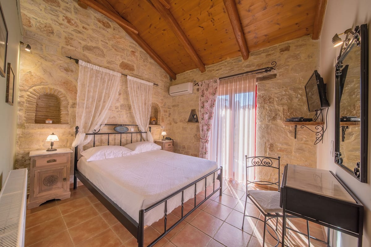 A bedroom with stone walls, a wooden ceiling, a bed with white linens, a nightstand with a lamp, a desk with a mirror, and a chair.
