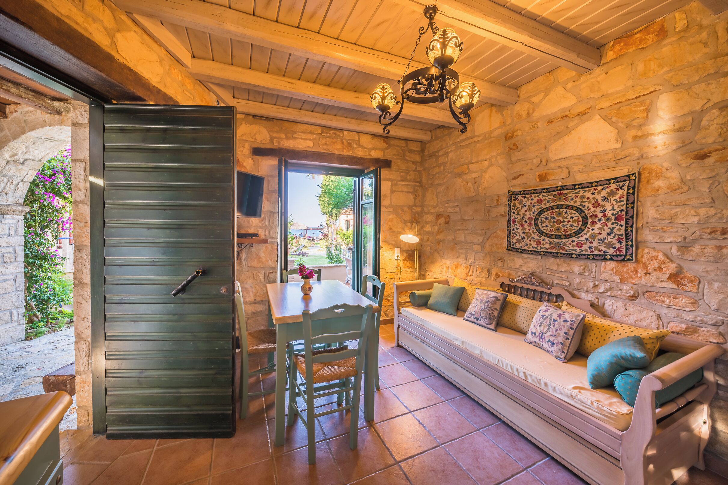 A cozy room with a stone wall, a wooden ceiling, a dining table with chairs, a sofa with cushions, and a chandelier.