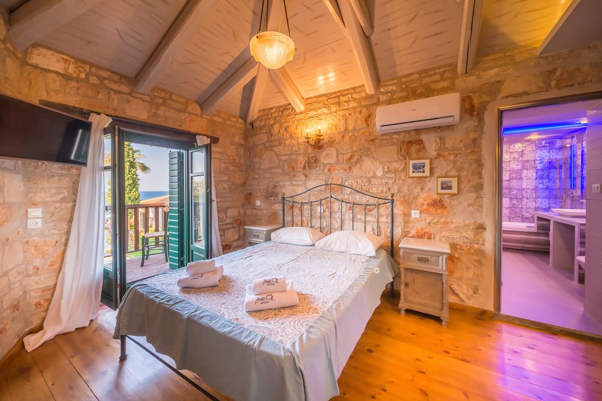 A bedroom with a stone wall, a bed with white linens, a nightstand, and an open door leading to a balcony.