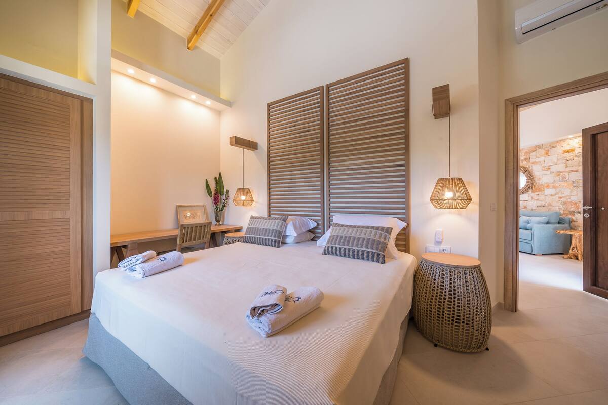 A modern bedroom with a large bed, bedside tables, a wall-mounted lamp, and a view into another room.