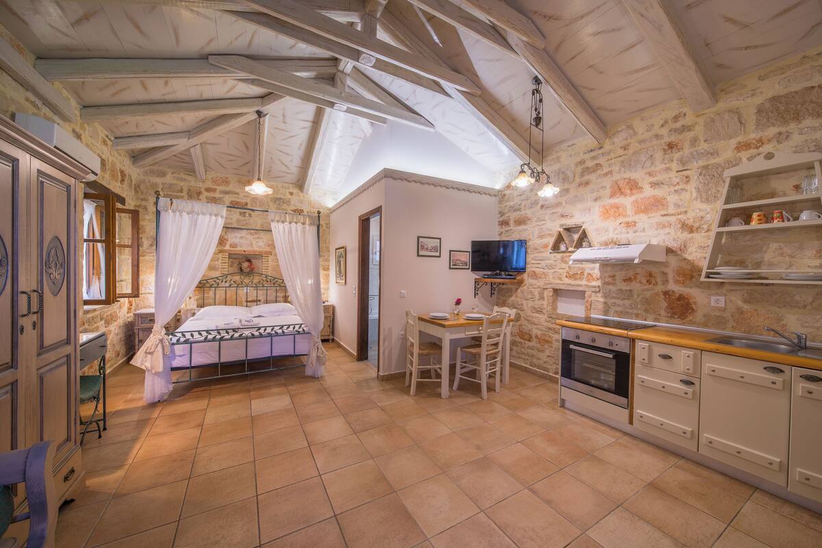 A bedroom with a bed, a bedside table, a window with curtains, a stone wall, a kitchen area with a stove, and a sink.