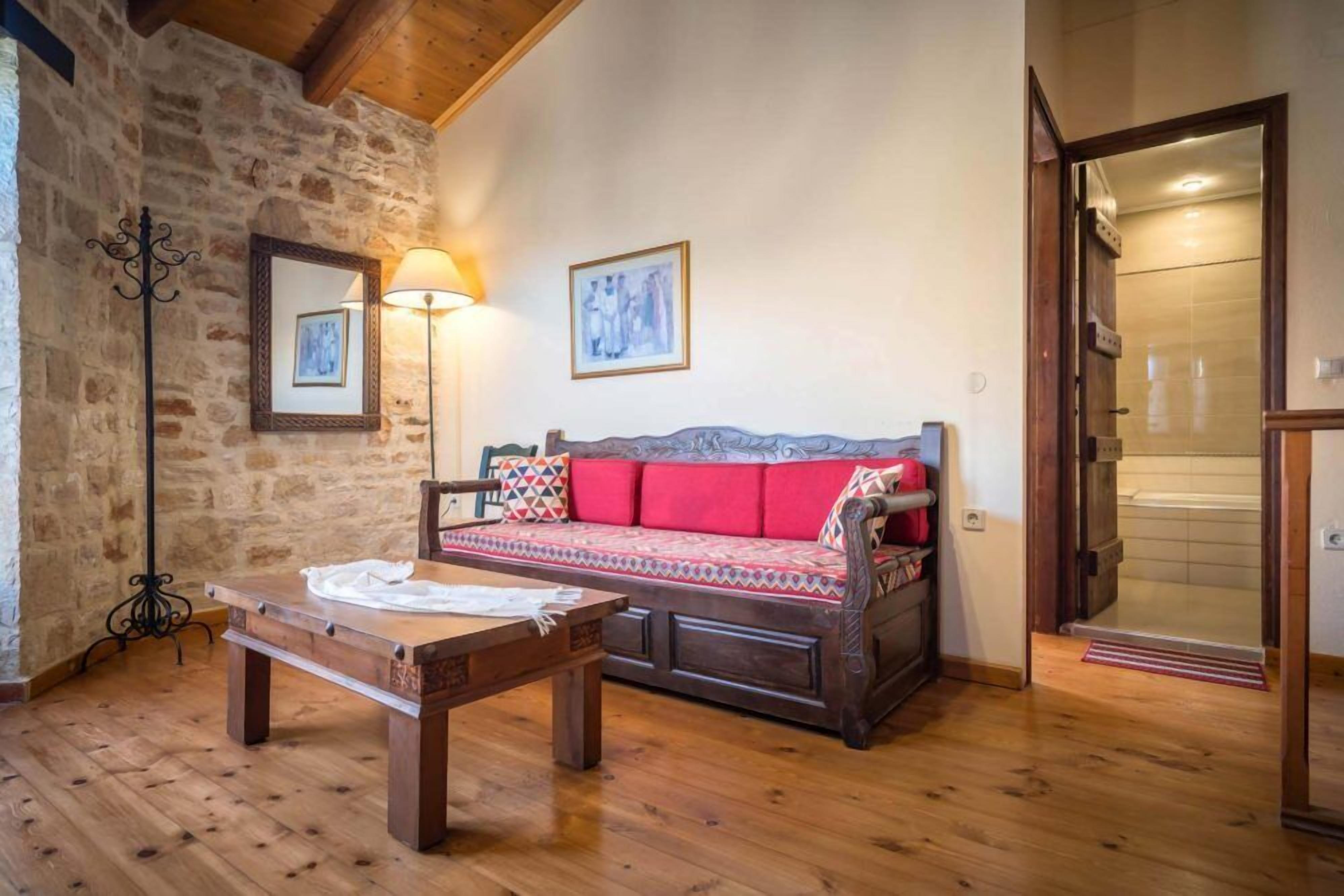 A cozy room with a wooden table, a sofa with red cushions, a stone wall, and a framed picture on the wall.
