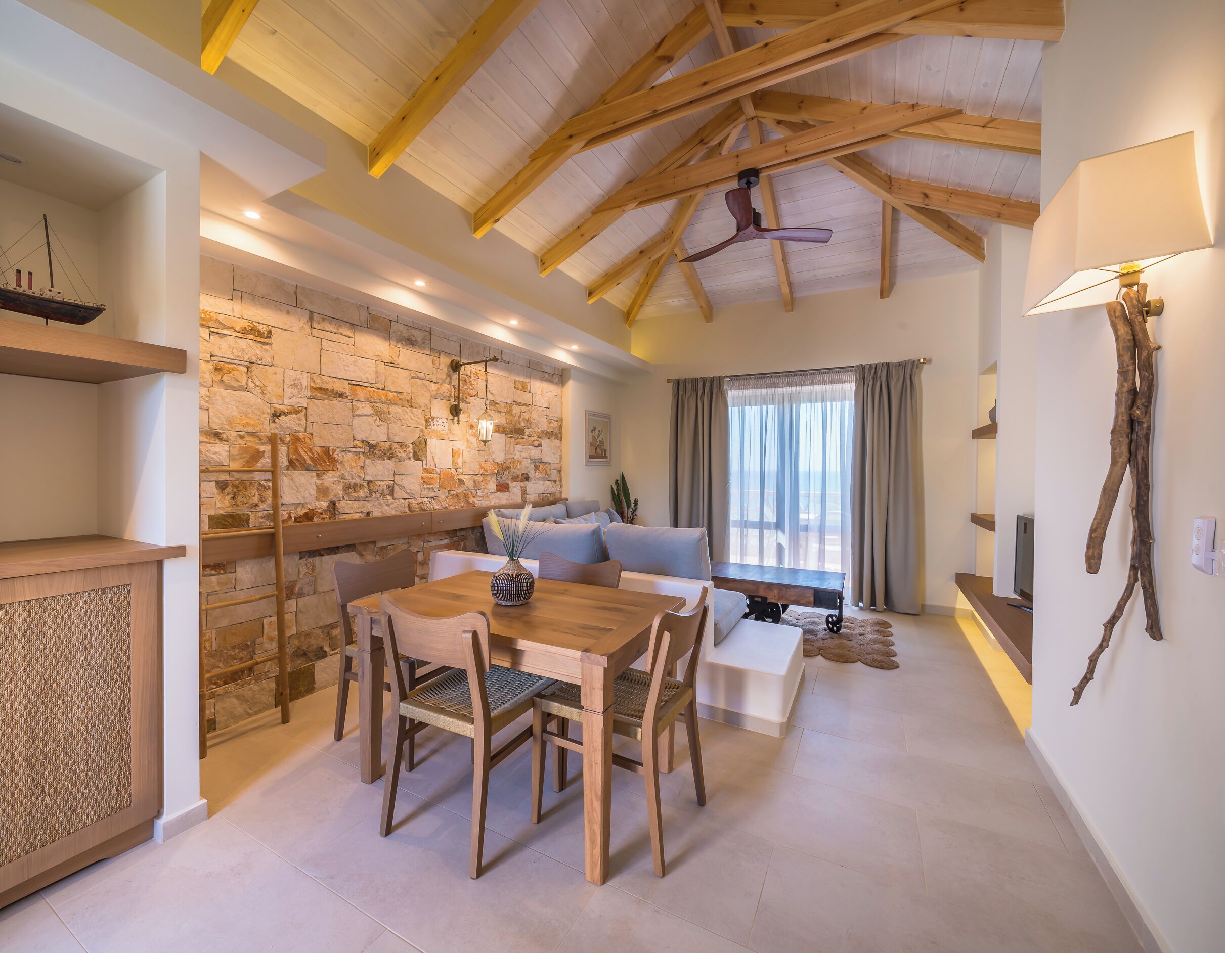 A spacious living area with a wooden ceiling, stone wall, and a dining table with chairs.