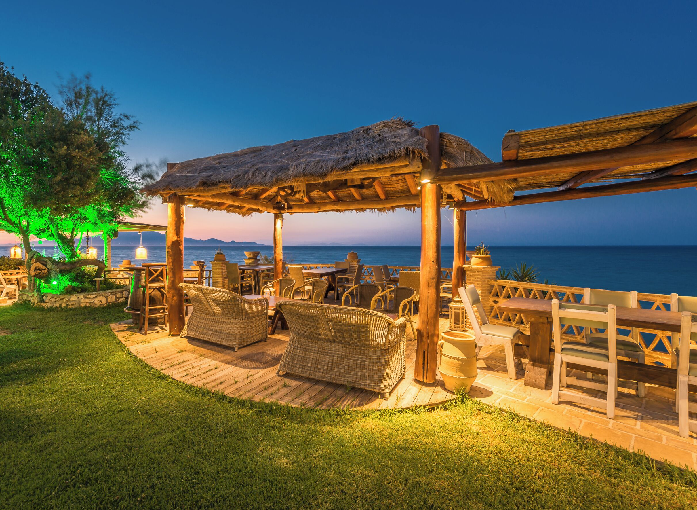 An outdoor seating area with wicker furniture and a thatched roof, overlooking a body of water.
