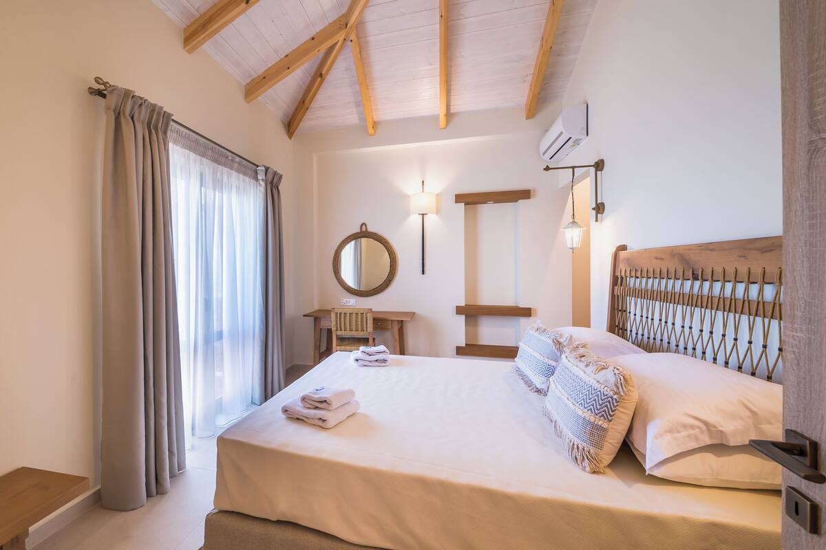 A bedroom with a wooden ceiling, a bed with white linens, a bedside table, a mirror, and a wall-mounted lamp.