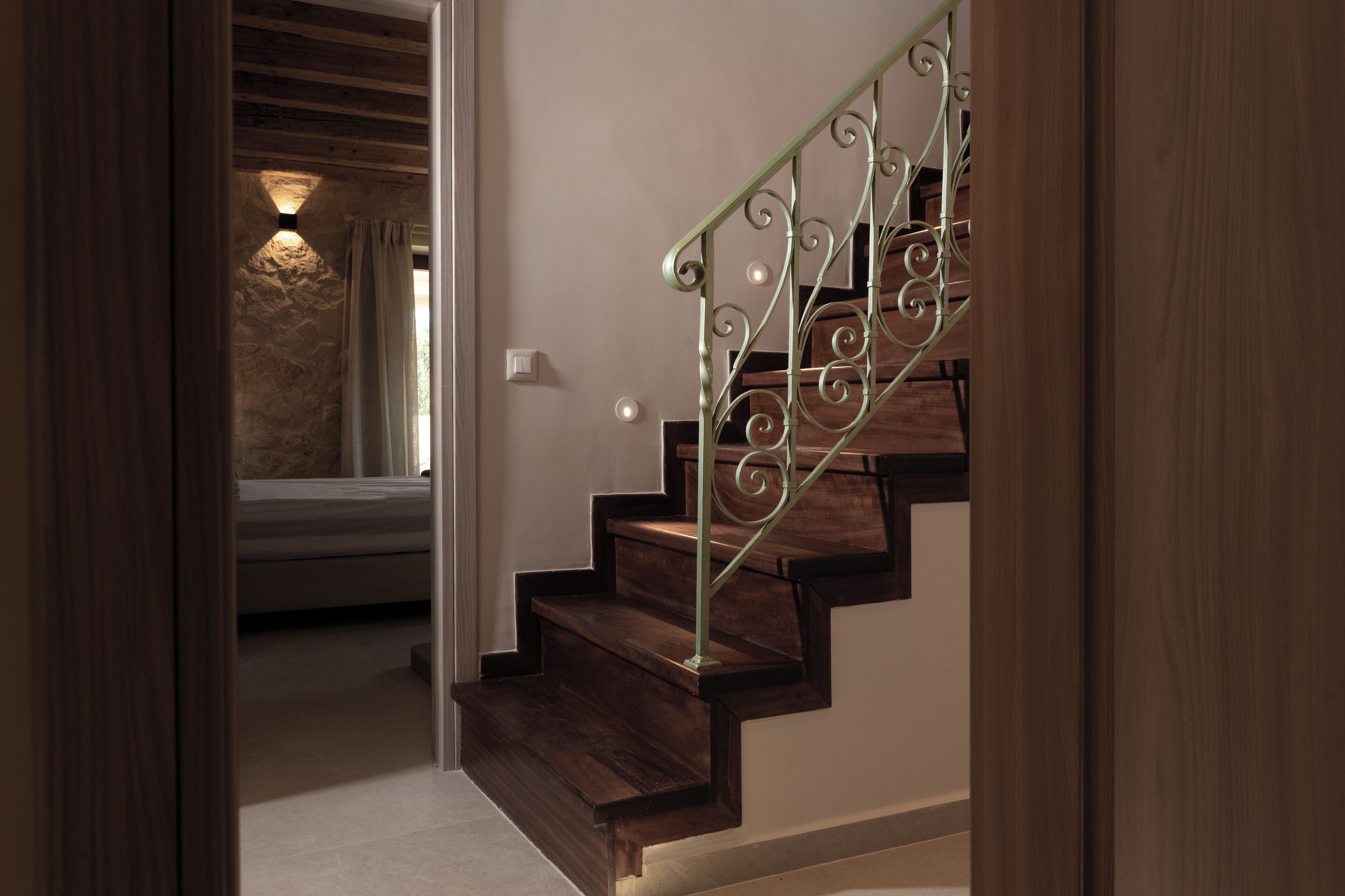 Escada de madeira com corrimão de metal que leva a um quarto com cama e luminária fixada na parede.