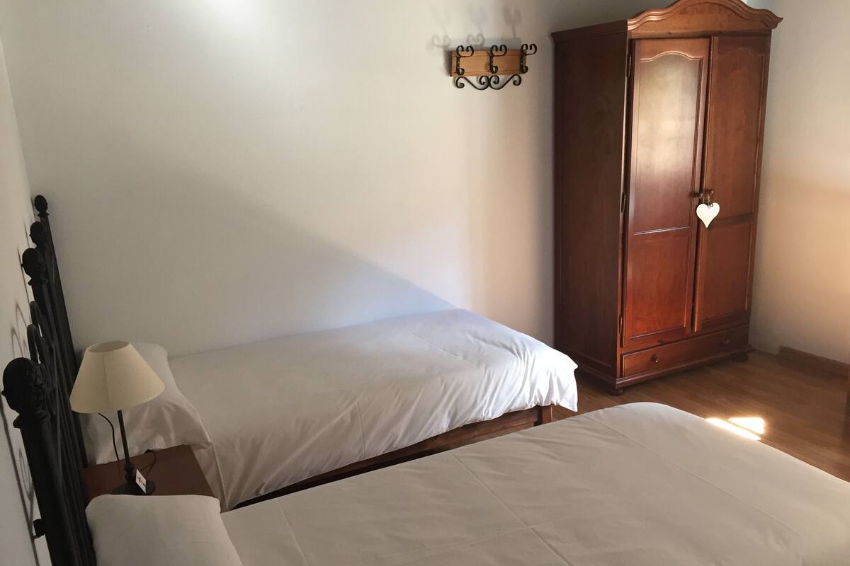 Quarto com duas camas de solteiro, um guarda-roupa de madeira e uma luminária fixada na parede.