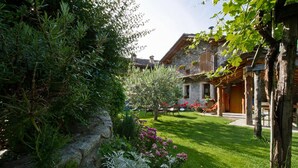 Ein Steinhaus mit einem hölzernen Balkon, umgeben von einem gepflegten Garten mit verschiedenen Pflanzen und Blumen.