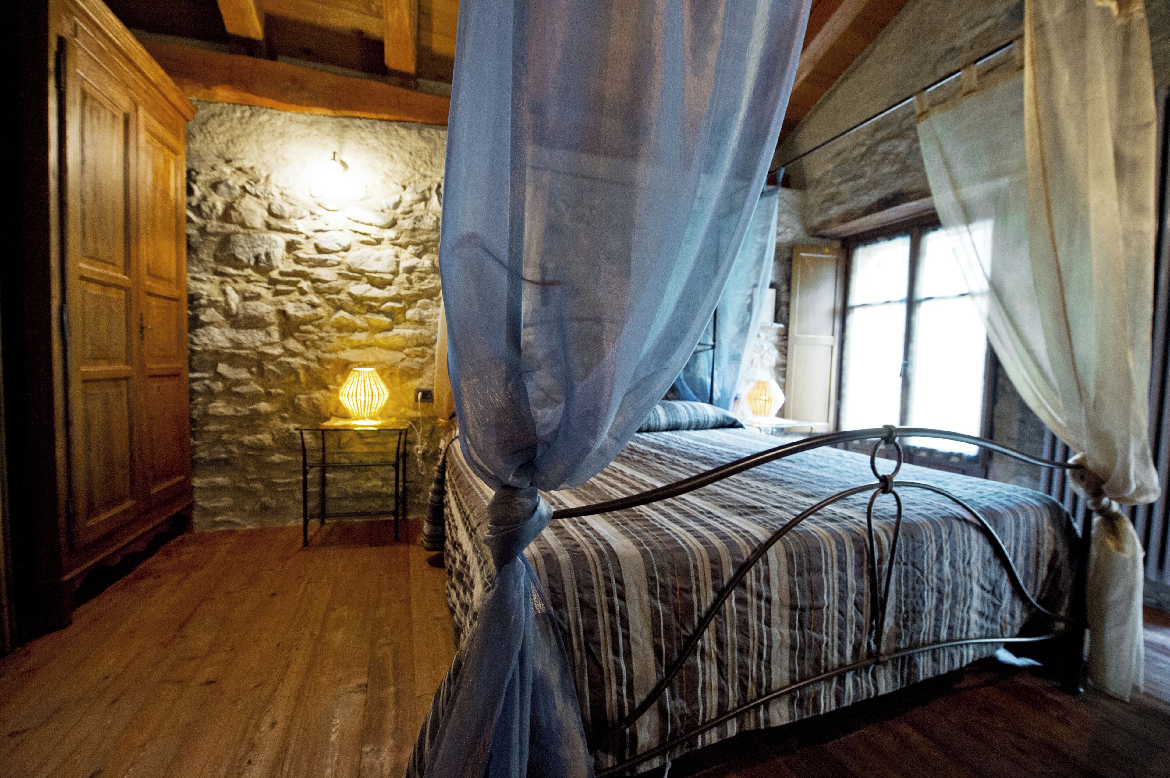 Un dormitorio con pared de piedra, una cama con estructura metálica, suelo de madera y una mesita de noche con lámpara.