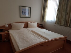 A double bed with white linens, a bedside table with a lamp, a framed picture on the wall, and a window with curtains. - Ratskeller Nörten (Norten-Hardenberg)