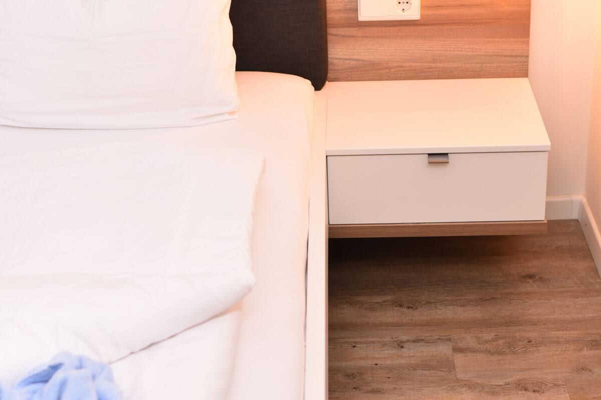 A neatly made bed with white linens and a blue towel, a wooden headboard, a bedside table with a lamp, and a wooden floor.