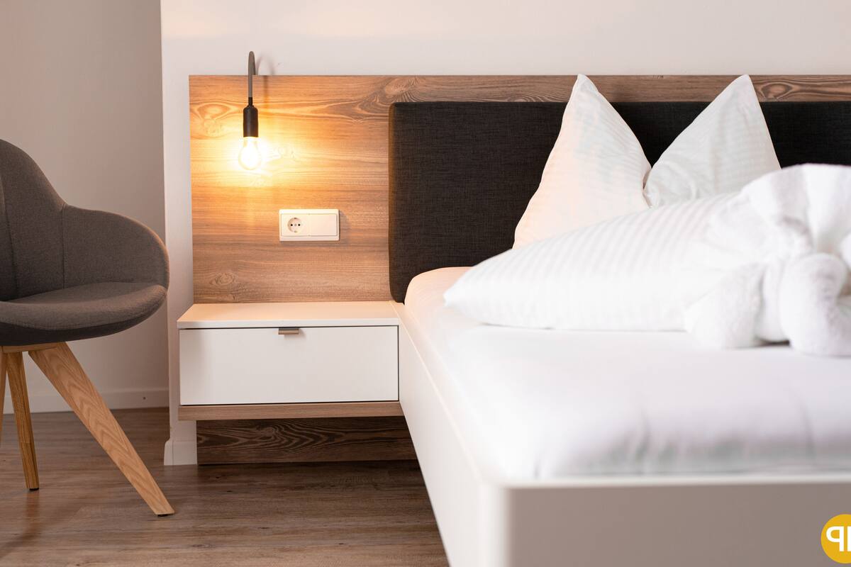 A modern bedroom with a wooden headboard, a bedside table with a lamp, a white bed, and a grey chair.