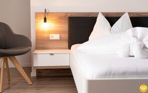 A modern bedroom with a wooden headboard, a bedside table with a lamp, a white bed, and a grey chair. - Pension Pernthaler (Silandro)