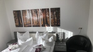 A hotel room with a bed, pillows, a bedside table with a lamp, a chair, and a wall-mounted painting of a forest. - Gästehaus Birke (Titisee-Neustadt)