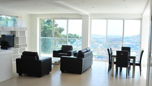 Una sala de estar moderna con un amplio ventanal, sofás negros, una mesa de comedor con sillas y un televisor en un estante blanco.