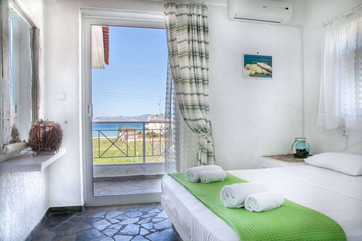 Habitación de hotel con una cama, una toalla verde, una mesita con un florero y vistas al mar a través de una puerta.