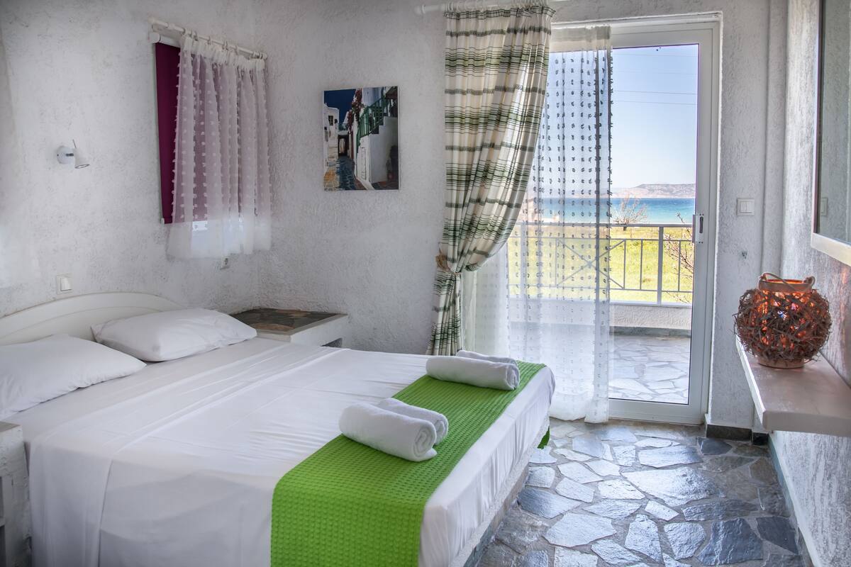 Un dormitorio con cama, ventana con cortinas, vista al mar y un jarrón decorativo.