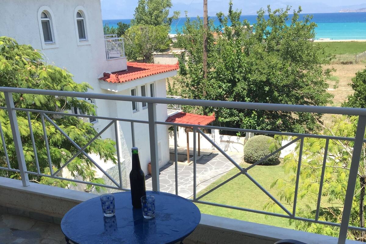 Un balcón con una mesa redonda y dos sillas, con vista a la costa y al mar, y un edificio de techo rojo.
