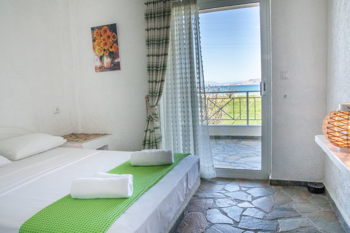 Habitación de hotel con cama, un tapete verde, un cuadro de girasoles, balcón con vistas y suelo de piedra.