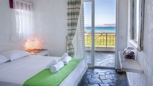 A bedroom with a bed, a nightstand, a green blanket, a window with curtains, and a view of the sea. - Phoenix Beachside Condos (Velo-Vocha)
