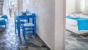 A room with a stone floor, a blue table with chairs, a bed with a blue bedspread, and a view of the sea through a window. - Phoenix Beachside Condos (Velo-Vocha)