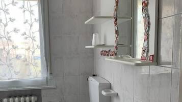 Ein Badezimmer mit Toilette, Waschbecken und Spiegel. Es gibt ein Fenster mit Jalousien, eine Heizung und einen Handtuchhalter.