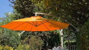 Un patio al aire libre con sillas y mesas de plástico blanco, un paraguas naranja y plantas en macetas.